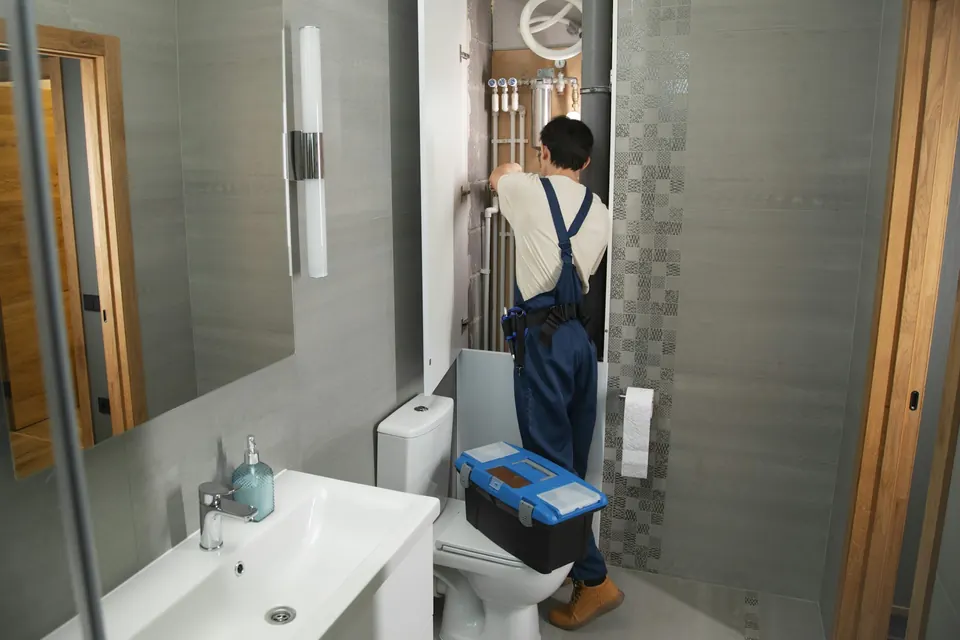 A tradesperson in navy overalls working on exposed pipe work behind a bathroom wall, toolbox open on the floor