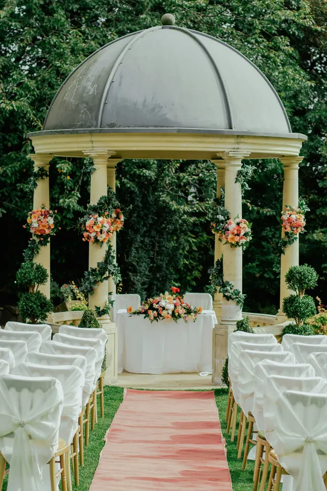 A garden pavilion set for a ceremony, a top table of peach and cream flowers between stone columns