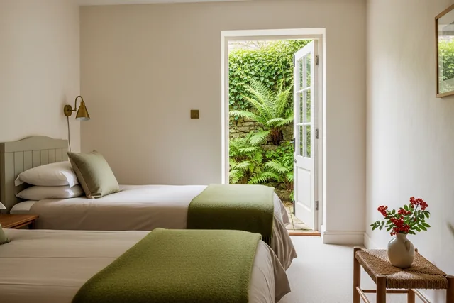 A small ground-floor room with two single beds, moss green blankets and a glazed door open onto a walled fern garden
